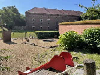 Ein Spielplatz mit Rutschbahnen und einem großen, roten Sandkasten. Im Hintergrund ist ein historisches Gebäude zu sehen, umgeben von Bäumen und Grün.