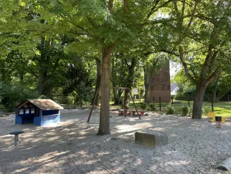 Ein Spielplatz mit einem Sandbereich und verschiedenen Spielgeräten. Umgeben von großen Bäumen und Grünfläche.