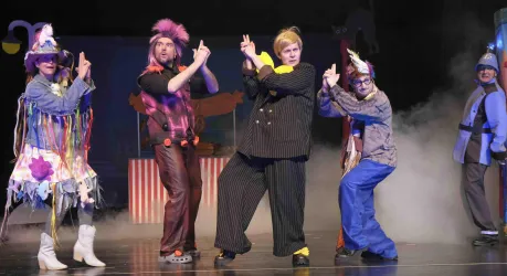 Eine Gruppe von fünf Schauspielern in bunten Kostümen steht auf einer Bühne. Sie posieren mit lustigen Gesten und wirken fröhlich und energiegeladen.