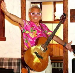 Ein fröhlicher Mann in einem bunten Hemd mit einem Akustikgitarren steht in einem gemütlichen Raum. Er lächelt und scheint bereit zu musizieren.