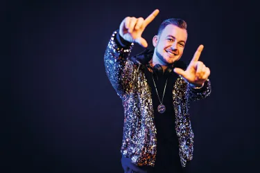 Ein junger Mann trägt eine glitzernde Jacke und Kopfhörer. Er lächelt und macht mit seinen Händen ein Zeichen.