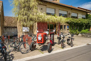 Ein historischer Traktor steht vor einem Gebäude, umgeben von Fahrrädern. Die Fassade ist grün bewachsen und die Sonne scheint auf die Szene.