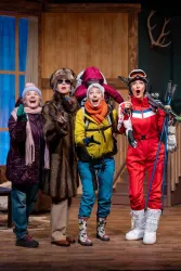 Vier Frauen in auffälligen Skihosen und Winterbekleidung stehen begeistert in einem Raum. Sie scheinen Spaß zu haben und lächeln fröhlich in die Kamera.