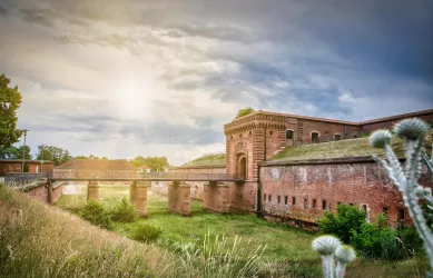Eine alte Festung mit roten Ziegelmauern und einem kleinen Brückenübergang. Im Hintergrund leuchtet die Sonne durch einen bewölkten Himmel.