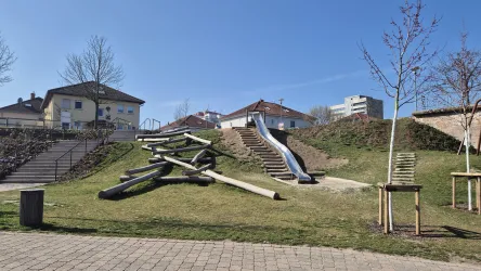 Ein Spielplatz mit Rutschen und Holzspielgeräten auf einer grünen Wiese. Im Hintergrund sind Wohnhäuser und ein blauer Himmel zu sehen.