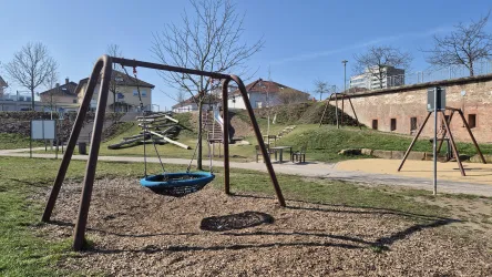 Ein Spielplatz mit einer Schaukel und einem großen Sandbereich. Im Hintergrund sind einige Bäume und Wohngebäude zu sehen.