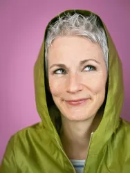 Eine Frau mit grauen Haaren trägt eine grüne Jacke mit Kapuze. Sie hat ein freundliches Lächeln und einen verspielten Blick vor einem rosa Hintergrund.