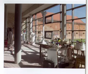 Ein helles Atelier mit großen Fenstern und einem Blick auf eine Backsteinmauer. Auf einem Tisch sind verschiedene Werkzeuge und Materialien verteilt.