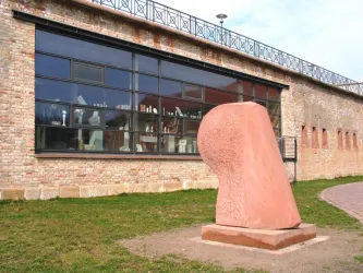 Eine moderne Skulptur aus rotem Material steht vor einem murartigen Gebäude. Im Hintergrund sind große Fenster, durch die verschiedene Kunstwerke zu sehen sind.