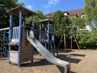 Ein Spielplatz mit einer Rutsche und Schaukeln. Im Hintergrund sind Bäume und ein Haus zu sehen.