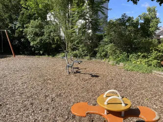 Ein Spielplatz mit einem Schaukelsitz und einem Wippgerät. Die Umgebung ist grün und sonnig, mit vielen Bäumen.