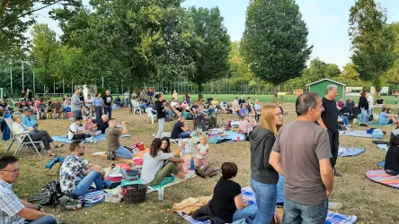 Eine große Menschenmenge im Freien, die auf Decken sitzt und sich unterhält. Bäume umrahmen den Ort und es wirkt entspannt und gesellig.