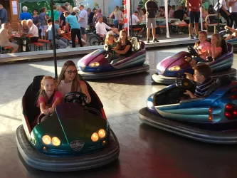 Eine Gruppe von Kindern fährt mit Autoscootern. Im Hintergrund sind viele Menschen zu sehen, die die Atmosphäre genießen.