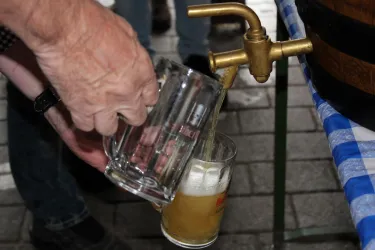 Ein Mensch gießt Bier aus einem Fass in ein Glas. Der Tisch ist mit einer karierten Tischdecke gedeckt.