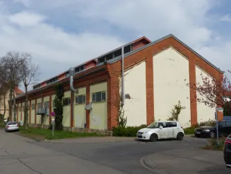 Ein großes, altes Gebäude mit roter Backsteinfassade und einem flachen Dach. Im Vordergrund stehen mehrere Autos und es gibt einige Bäume und Sträucher.