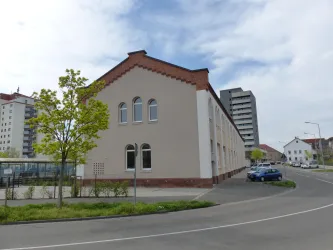 Ein mehrstöckiges Gebäude neben einem modernem Wohnblock ist in der Aufnahme zu sehen. Die Straße und einige grüne Bäume umgeben die Szene.