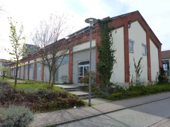 Ein modernes Gebäude mit einem gut gepflegten Außenbereich. Die Fassade ist teilweise mit Pflanzen bewachsen und es gibt Gehwege sowie Straßenbeleuchtung.