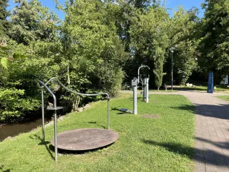 Ein grüner Park mit Fitnessgeräten im Freien. Bäume und ein kleiner Wasserlauf sind im Hintergrund zu sehen.