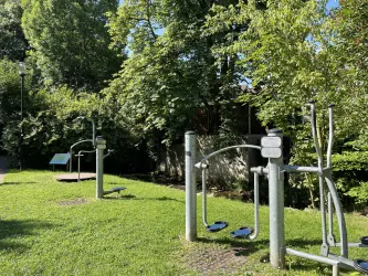 Ein Outdoor-Fitnessbereich mit mehreren Trainingsgeräten steht in einem grünen Park. Umgeben von Bäumen bietet er einen einladenden Platz für Sport und Bewegung.