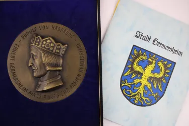 Eine Medaille mit dem Abbild von Rudolf von Habsburg und einer historischen Inschrift. Daneben liegt eine Broschüre mit dem Wappen der Stadt Germersheim.
