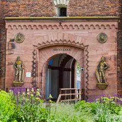 Ein historischer Torbogen mit zwei stehenden Kriegerstatuen. Umgeben von bunten Blumen und grüner Vegetation.