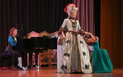 Eine Theateraufführung mit Schauspielerinnen in historischen Kostümen. Eine Pianistin spielt am Klavier, während eine Darstellerin auf der Bühne agiert.