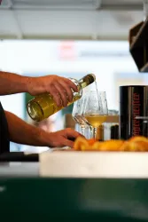 Eine Hand gießt ein Getränk aus einer Flasche in zwei Gläser. Im Vordergrund sind einige Croissants zu sehen.
