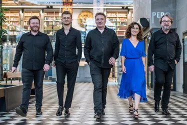 Fünf Personen gehen in einem modernen Raum. Vier von ihnen tragen schwarze Kleidung, während die Frau ein blaues Kleid anhat.