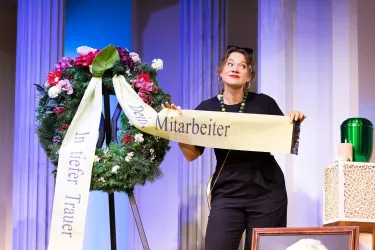 Eine Schauspielerin zeigt ein Trauergesteck mit einem Banner. Sie steht im Mittelpunkt der Bühne und wirkt nachdenklich.