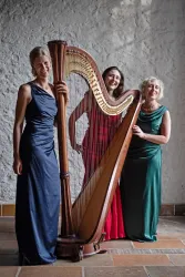 Drei Frauen stehen lächelnd vor einer Harfe. Sie tragen elegante Kleider in verschiedenen Farben.