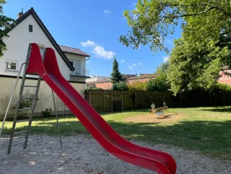 Ein Spielplatz mit einer roten Rutsche und viel Grün. Im Hintergrund ist ein Haus zu sehen.