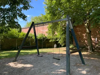 Eine Schaukel auf einem Spielplatz mit sandigem Boden. Im Hintergrund sind Bäume und eine Mauer zu sehen.