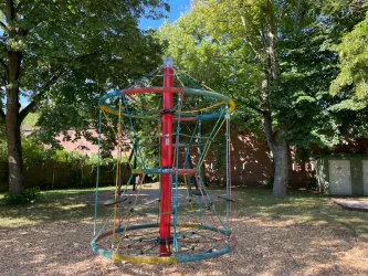 Ein Spielplatz mit einem bunten Klettergerät. Umgeben von grünen Bäumen und Sonnenlicht.