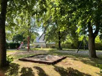 Ein schöner Spielplatz mit einer Rutsche und einer Schaukel in einem grünen Garten. Überall stehen große Bäume und viel Sonnenlicht scheint hindurch.