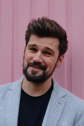 Ein junger Mann mit einem lächelnden Gesicht steht vor einer pinken Wand. Er trägt einen grauen Blazer und ein schwarzes T-Shirt.