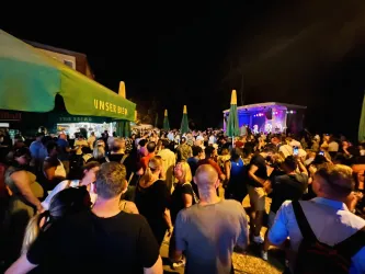 Eine große Menschenmenge versammelt sich bei einem Fest im Freien bei Nacht. Auf der Bühne sind Lichter und eine Band zu sehen, die Musik spielt.