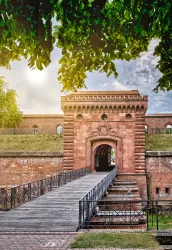 Ein historisches Tor mit einer Holzbrücke führt in eine Festungsanlage. Die Umgebung ist grün und sonnig, was eine einladende Atmosphäre schafft.