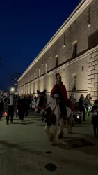 Eine Gruppe von Menschen mit einem Ritt auf einem Pferd. Der Reiter trägt einen roten Umhang und die Szene spielt sich in der Dämmerung vor einer historischen Mauer ab.