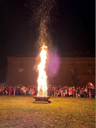 Ein großes Feuer brennt in der Nacht, umgeben von einer Menschenmenge. Im Hintergrund ist ein historisches Gebäude zu sehen.