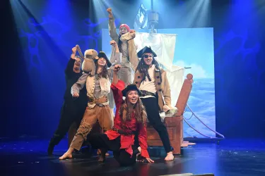 Eine Theateraufführung mit Piratenkostümen und einer hölzernen Schiffskonstruktion. Die Darsteller posieren fröhlich auf der Bühne.