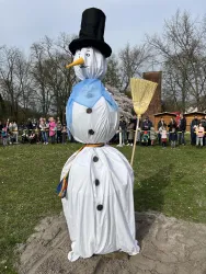Ein großer Schneemann mit einem schwarzen Zylinder und einem blauen Schal steht in einem Park. Im Hintergrund sind viele Menschen zu sehen, die zuschauen.