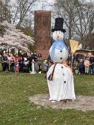Eine große, festliche Schneemanfigur steht in einem Park. Um ihn herum versammelt sich eine fröhliche Menge, darunter viele Kinder.