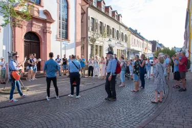 Eine Gruppe von Menschen versammelt sich in einer malerischen Straße, während ein Musiker auf der Gitarre spielt. Die Atmosphäre ist lebhaft und freundlich.