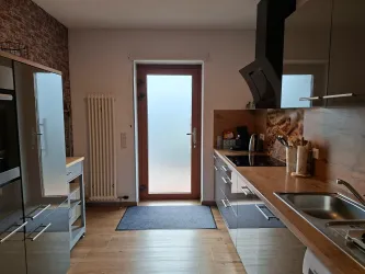 Eine moderne Küche mit Holzarbeitsplatte und glänzenden Schränken. Der Blick führt zur gläsernen Tür und einem hellen Eingang.