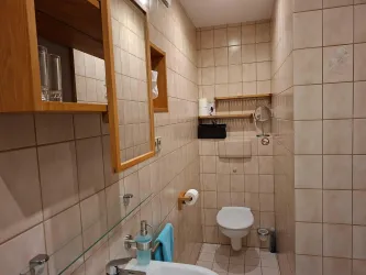 Ein Badezimmer mit hellen Fliesen, einem Waschbecken und einer Toilette. Holzregale und ein großer Spiegel ergänzen die Ausstattung.