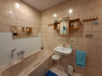 Ein modernes Badezimmer mit einer Badewanne und einem Waschbecken. Die Wände sind mit Fliesen in warmen Farbtönen dekoriert, und es gibt praktische Regale für Pflegeprodukte.