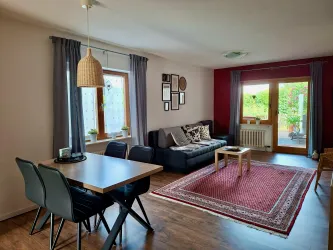 Ein gemütlicher Wohnraum mit einem Esstisch und Stühlen sowie einem Sofa. Helle Farben und große Fenster sorgen für eine einladende Atmosphäre.