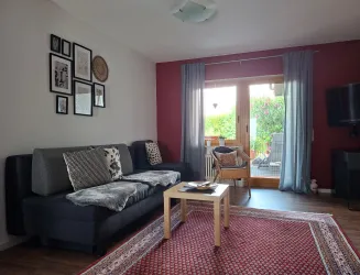 Ein gemütliches Wohnzimmer mit einem  Sofa und einem Holztisch. Große Fenster lassen viel Licht herein und bieten einen Blick auf den Garten.