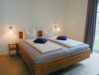 Ein helles Schlafzimmer mit zwei Einzelbetten und einer warmen Holzkommode. Die Wände sind hell gestrichen, und es gibt große Fenster mit Vorhängen.