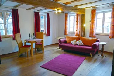 Ein gemütliches Wohnzimmer mit einem roten Sofa und einem kleinen Tisch. Große Fenster lassen viel Licht herein und die rot-orangen Vorhänge verleihen eine warme Atmosphäre.